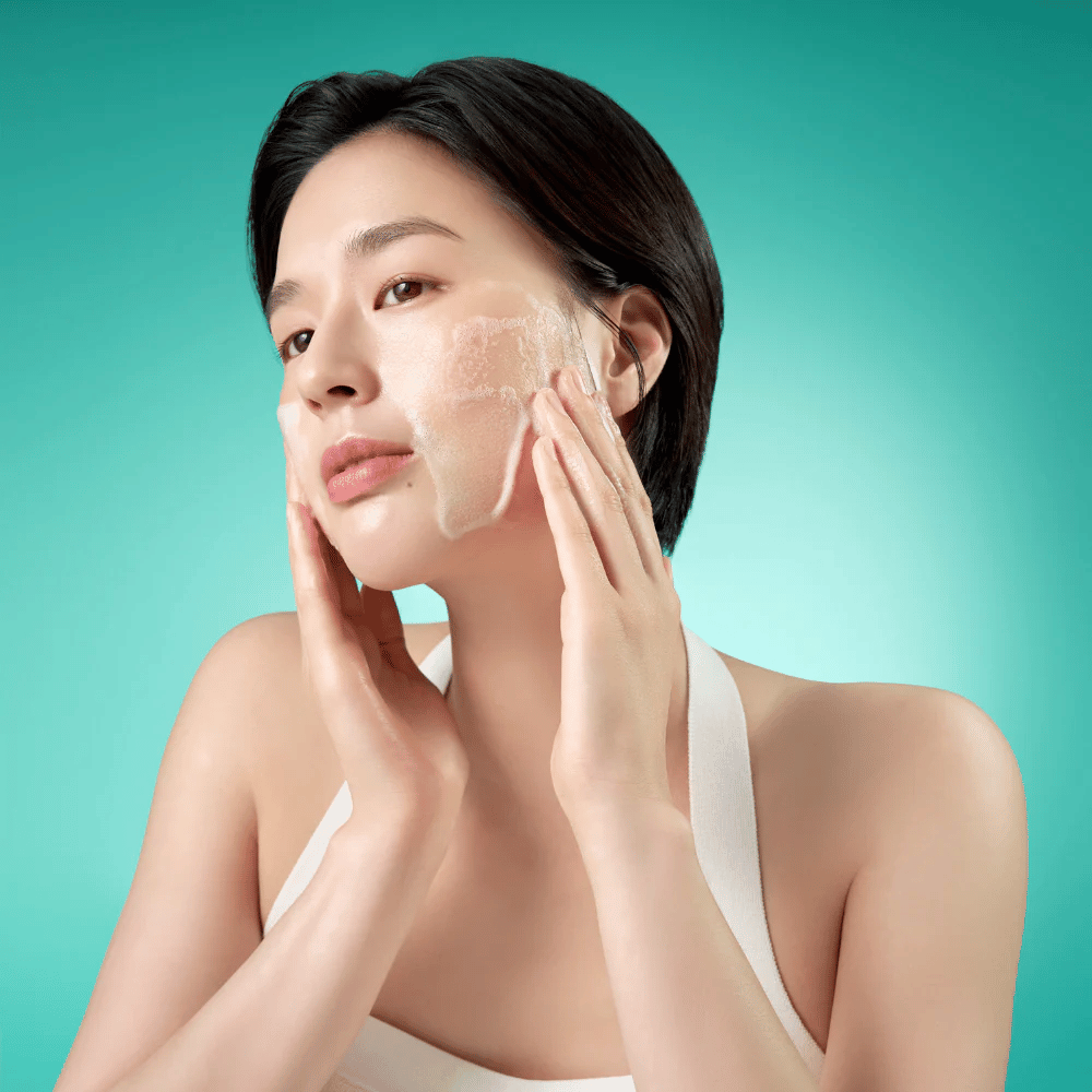 Mulher limpa o rosto com uma espuma de limpeza facial suave contra um fundo verde.