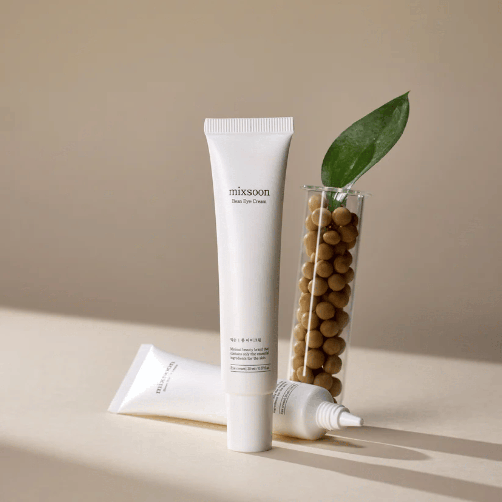 Dois tubos Mixsoon Bean Eye Cream colocados ao lado de um tubo de ensaio cheio de grãos de soja e uma folha de planta verde, sobre uma superfície bege com jogo de sombras.
