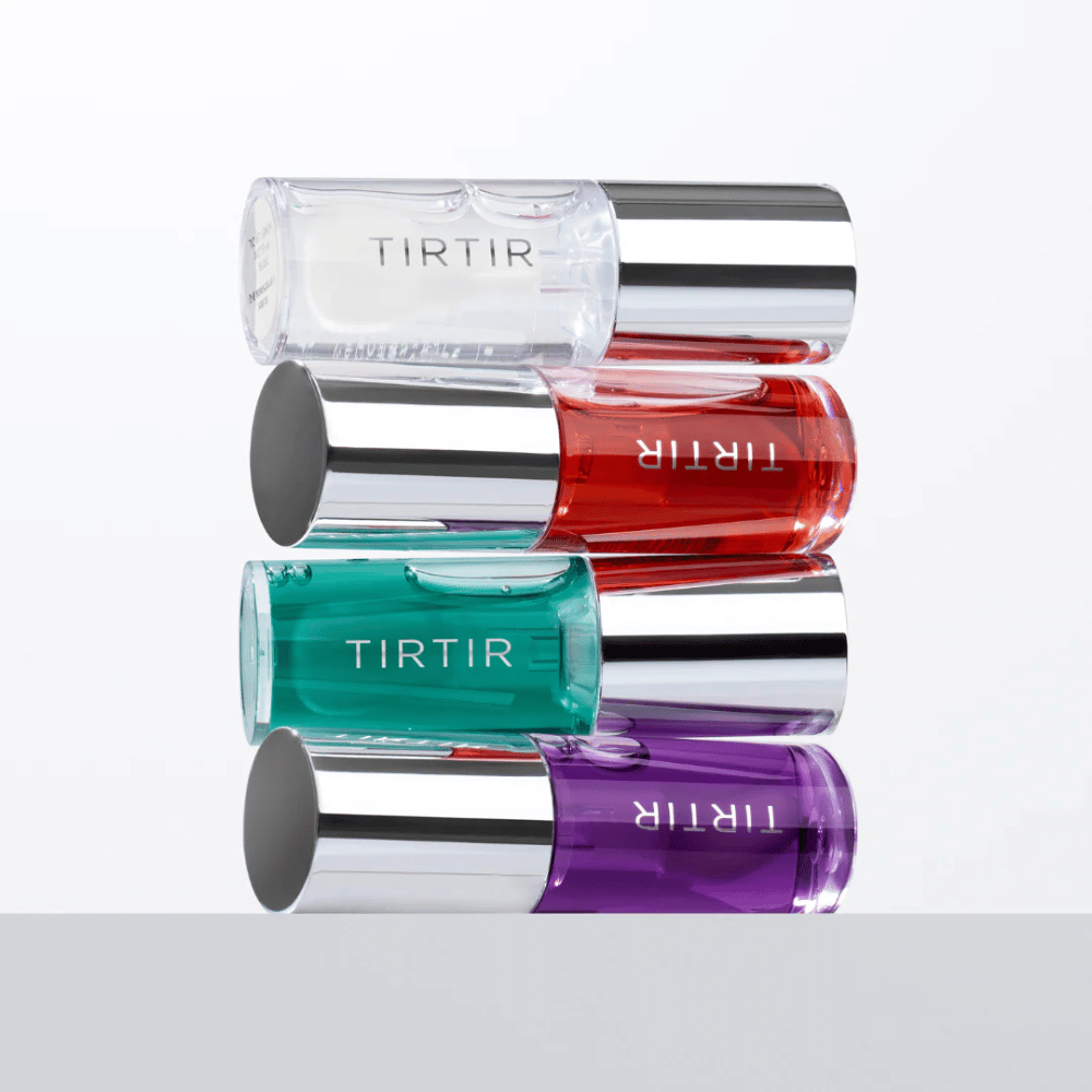 Quatro TIRTIR My Glow Lip Oil em diferentes cores – transparente, vermelho, verde e roxo – empilhados uns sobre os outros com tampas prateadas contra um fundo branco.