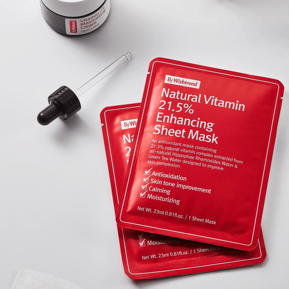 By Wishtrend Natural Vitamin 21.5% Enhancing Sheet mask em embalagens vermelhas, colocadas sobre uma superfície branca junto com um conta-gotas e um pote de creme facial.