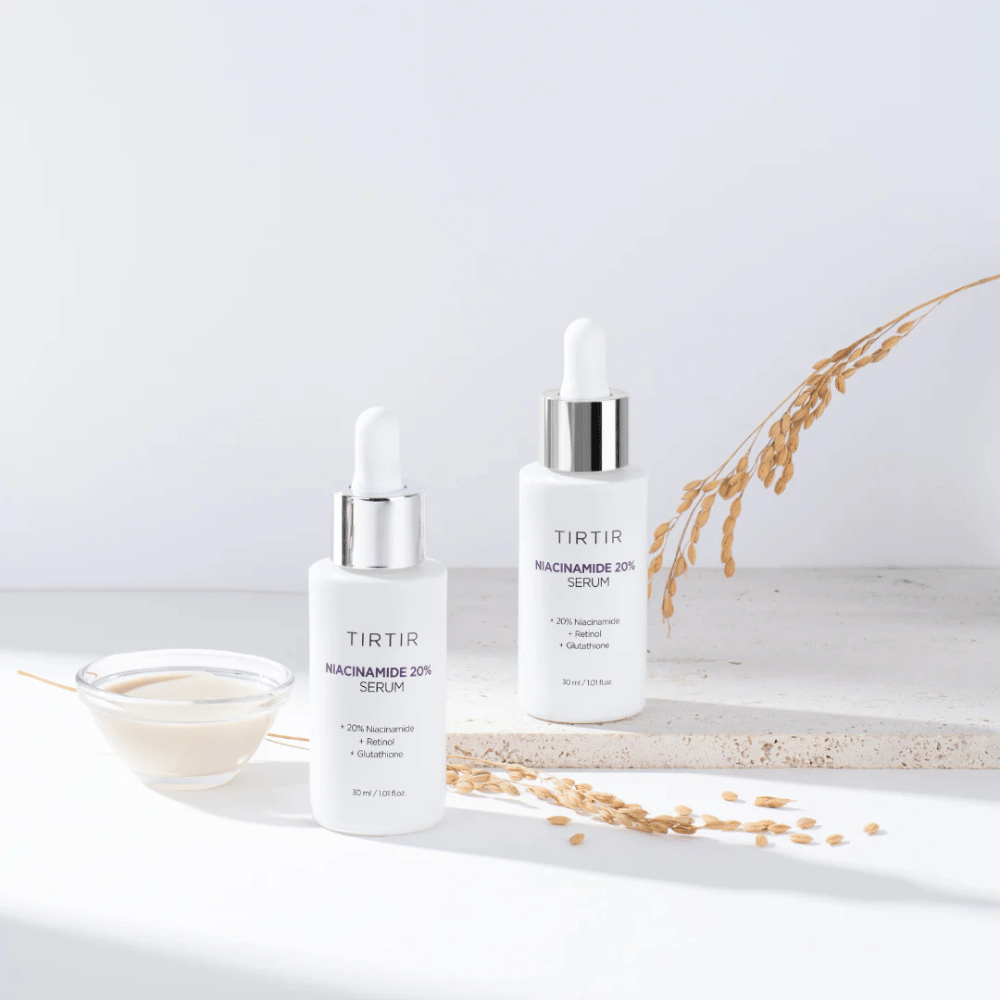 Duas garrafas de Tirtirs "Niacinamide 20% Serum" são exibidas sobre uma superfície clara junto com uma pequena tigela com uma substância cremosa e algumas hastes de plantas secas. O sérum contém 20% de niacinamida, retinol e glutationa, o que ajuda a melhorar a aparência e a textura da pele.