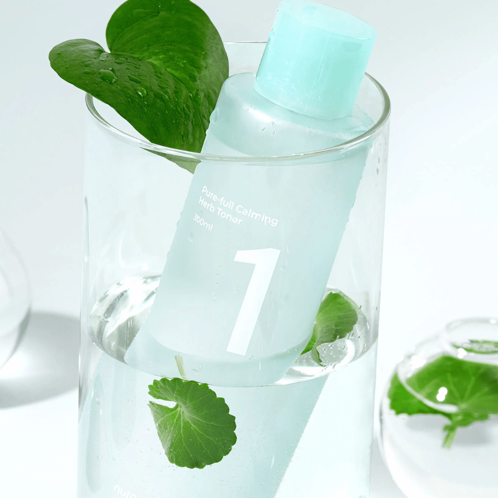 Uma garrafa com No.1 Pure-Full Calming Herb Toner colocada em um copo com água e folhas verdes. A garrafa tem uma tampa azul-claro e um rótulo com o texto "Pure-full Calming Herb Toner" e "300ml". O produto parece fresco com água e vegetação ao fundo, o que transmite uma sensação calmante e natural.