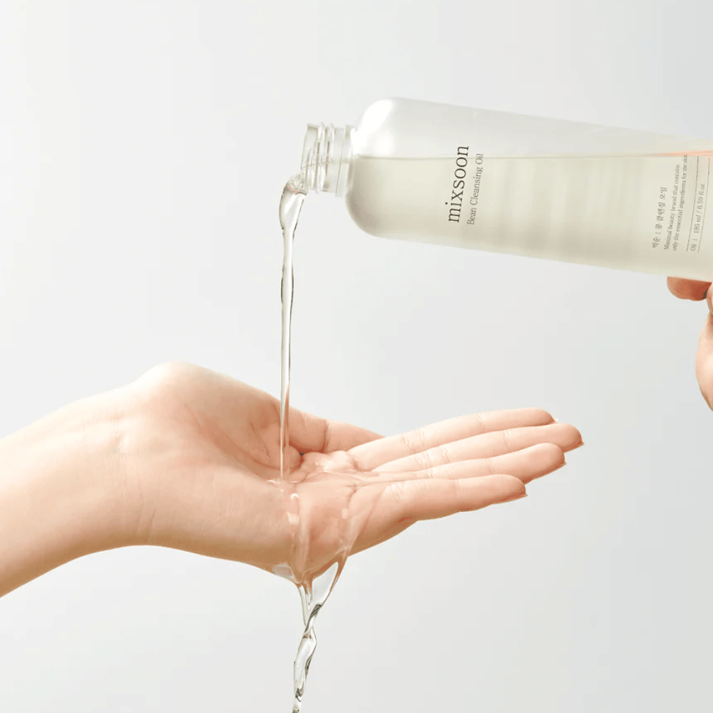Um óleo de limpeza claro é derramado de uma garrafa transparente com rótulo branco onde está escrito "MixSoon" em uma palma aberta. O óleo forma um jato brilhante entre a abertura da garrafa e a mão, demonstrando a consistência líquida do óleo. A mão está imóvel e posicionada de modo a recolher toda a quantidade de óleo de limpeza contra um fundo claro e uniforme.