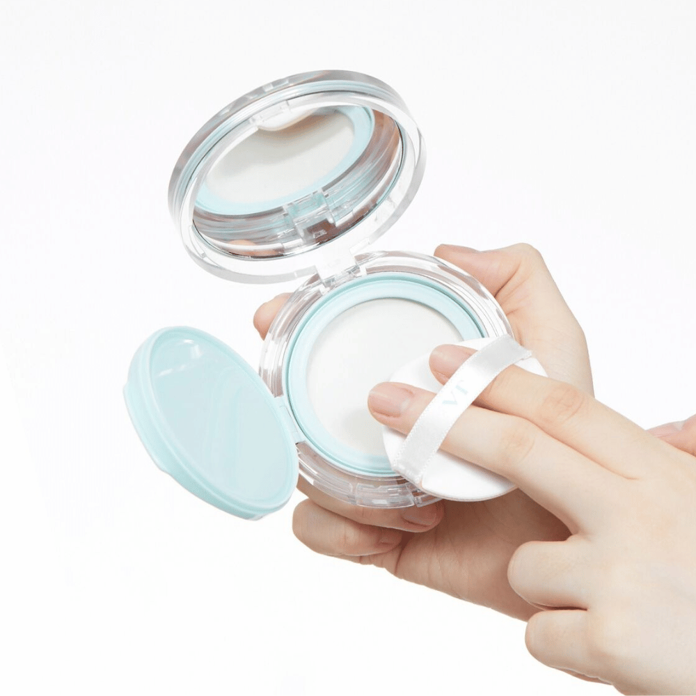 Uma mão segura uma pote aberta com VT PDRN Essence Glow Sun Pact. O aplicador está prestes a aplicar o pó, que tem uma superfície branca e uma borda azul ao redor do pote.