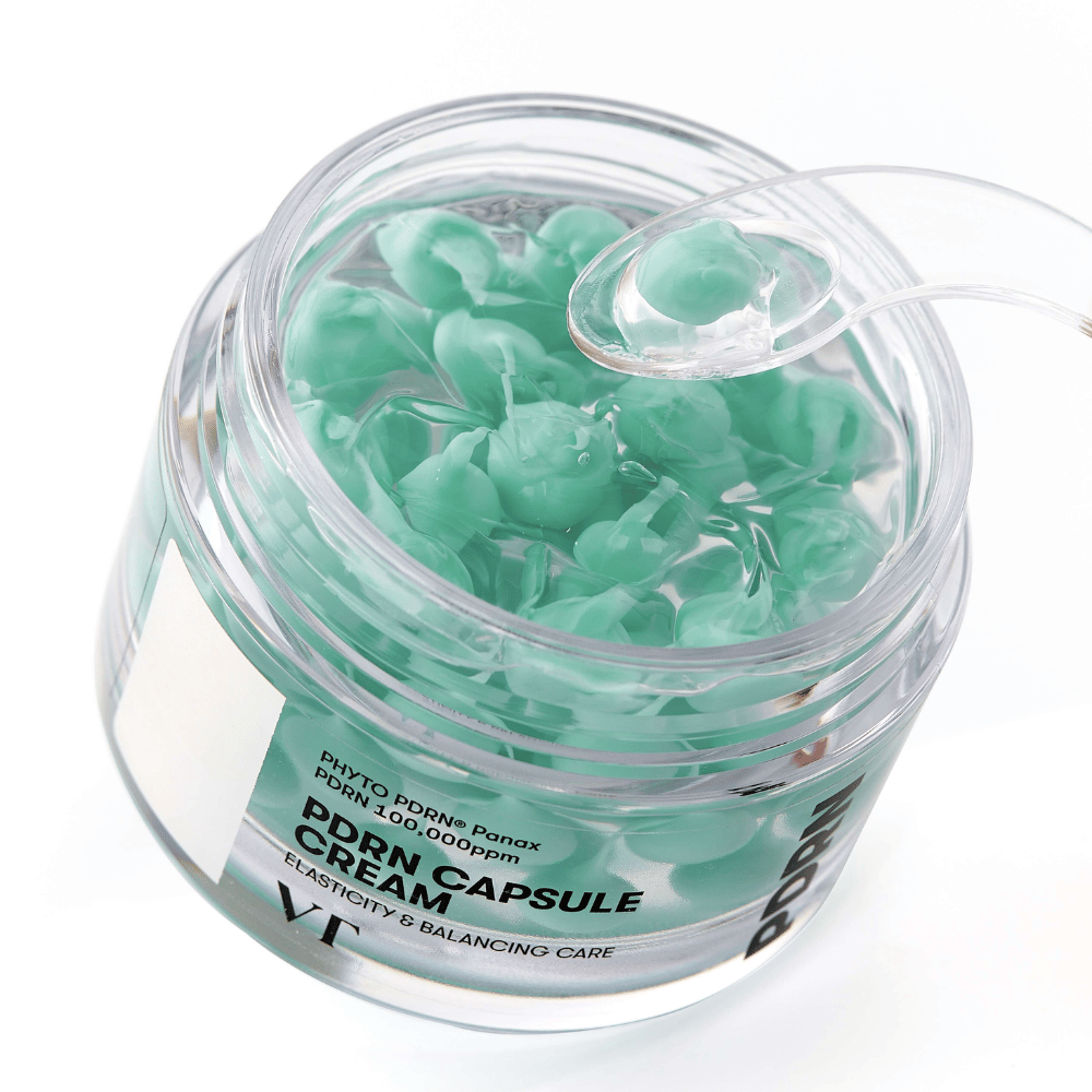 Close-up de um frasco aberto com VT PDRN Capsule Cream. O conteúdo consiste em pequenas cápsulas verdes em um gel transparente, com uma espátula incluída para aplicação. O texto no frasco diz "Elasticity & Balancing Care".