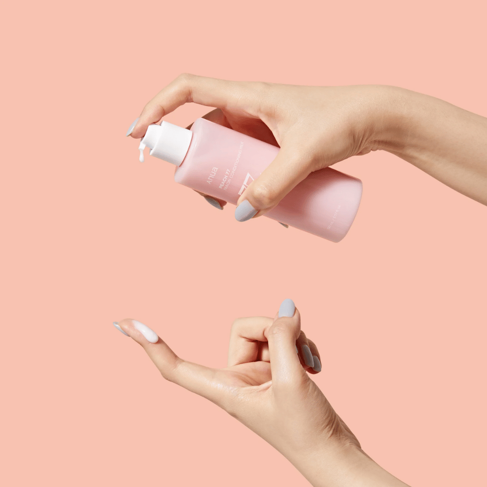A imagem mostra as mãos de alguém segurando uma garrafa com ANUA Peach 77% Niacin Conditioning Milk. Este produto de cuidados com a pele parece ter uma consistência leitosa e é projetado para cuidar e melhorar a condição da pele.