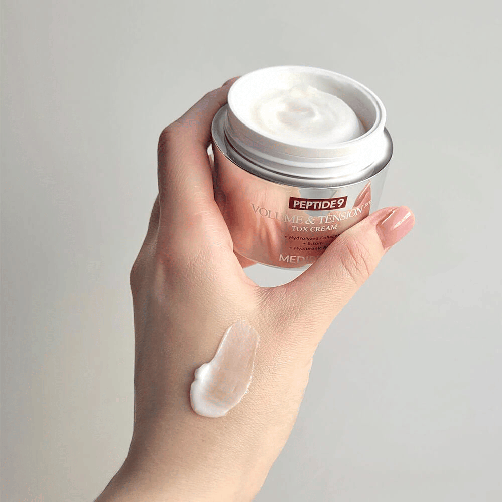 Mão segurando MEDI-PEEL Peptide 9 Volume & Tension Tox Cream com um pouco de creme no dorso da mão. A fórmula sedosa hidrata e dá firmeza e elasticidade à pele.