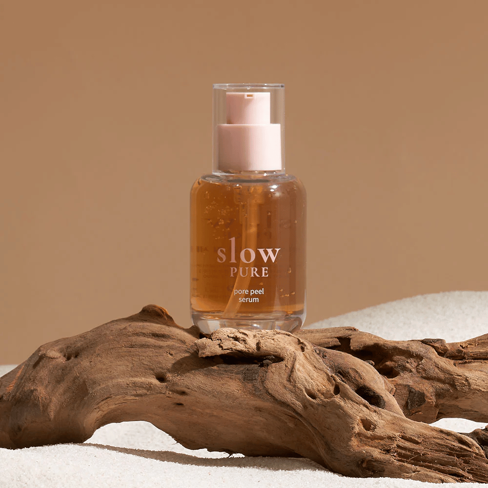 Slow Pure Pore Peel Serum colocado na areia com detalhe em madeira ao fundo, símbolo de cuidados naturais para a pele.