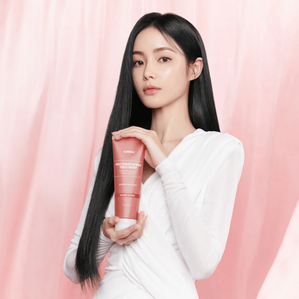 Mulher com cabelo longo e brilhante segurando KUNDAL Protein Bonding Treatment Violet Muguet contra um fundo rosa.