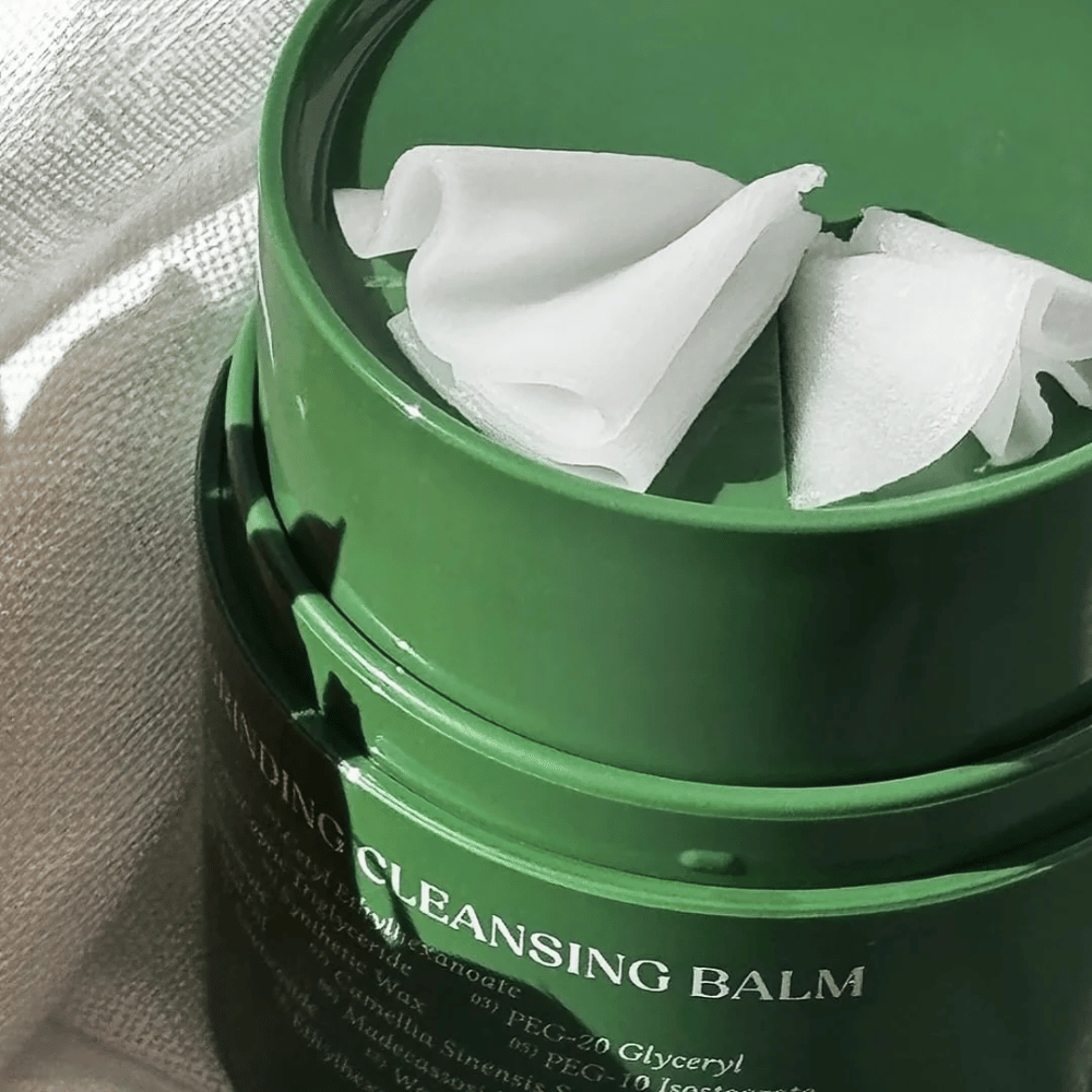 A imagem mostra um pote verde com "Cleansing Balm", que está aberto para revelar a espátula incluída e o conteúdo suave. A consistência cremosa reflete a luz, o que dá uma sensação de luxo e eficácia na limpeza.