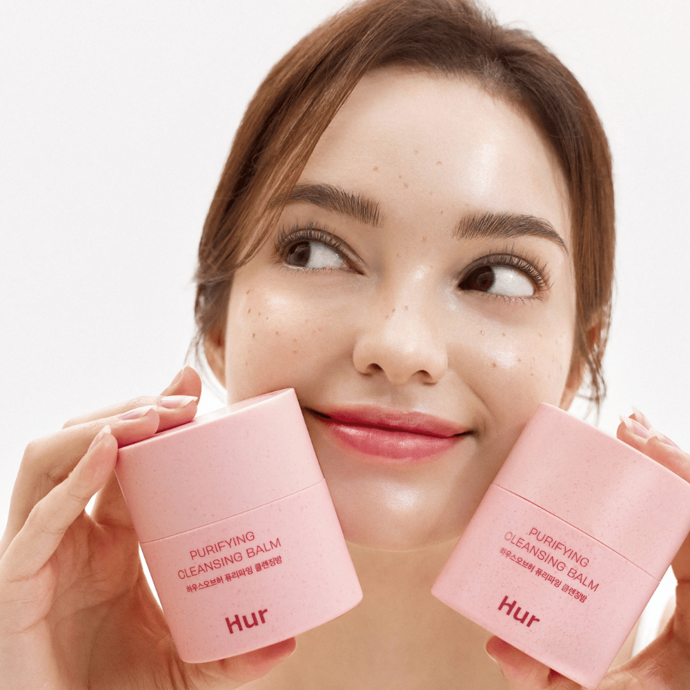 Modelo segurando dois potes Como Purifying Cleansing Balm. Bálsamo de limpeza suave que é adequado para pele sensível e proporciona uma sensação fresca e hidratada após a limpeza.
