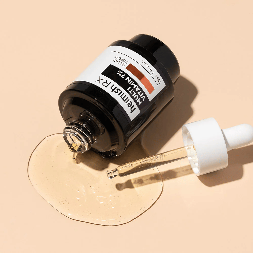 Heimish RX Multi Vitamina 7% Glow Serum deitado em um fundo bege com soro espalhado ao redor da garrafa e uma pipeta cheia de produto, destacando sua fórmula rica em nutrientes.