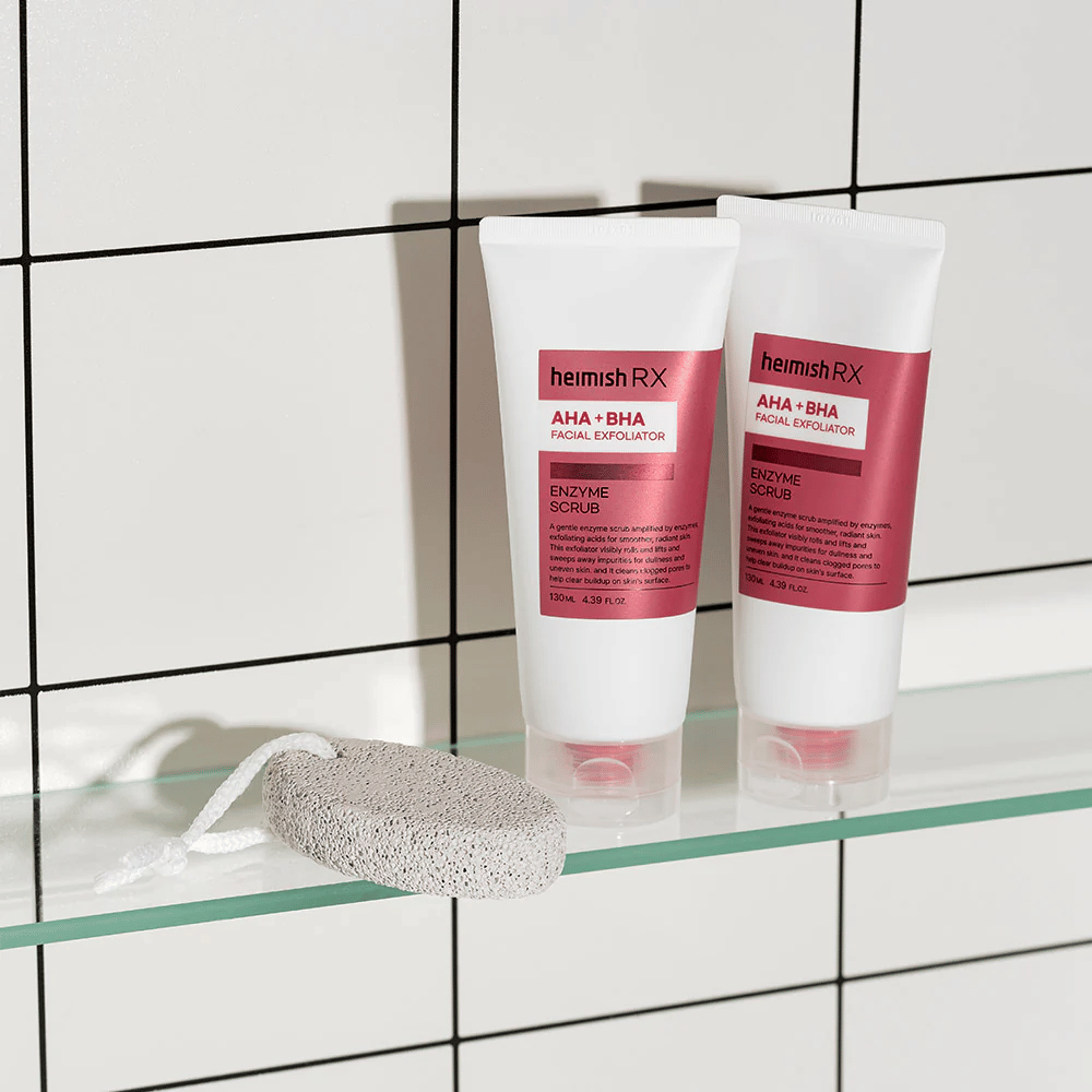Dois tubos Heimish RX AHA + BHA Facial Exfoliator colocados em uma prateleira de vidro em um banheiro com parede de azulejos brancos. Ao lado dos produtos está uma pedra-pomes com um cordão branco. Uma imagem de cuidados com a pele elegante e minimalista.