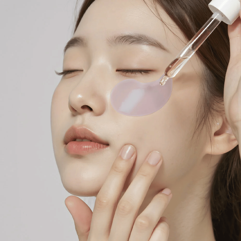 Uma mulher com um remendo ocular de hidrogel roxo aplica soro sob o olho com uma pipeta. O produto hidrata e cuida da pele sensível ao redor dos olhos.