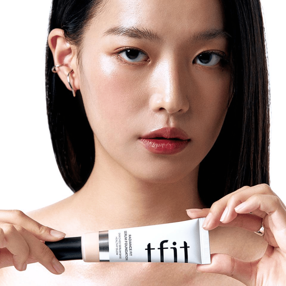Modelo com pele lisa e brilhante segurando TFIT Radiance Fit Serum Foundation, destacando o acabamento natural e o brilho do produto.