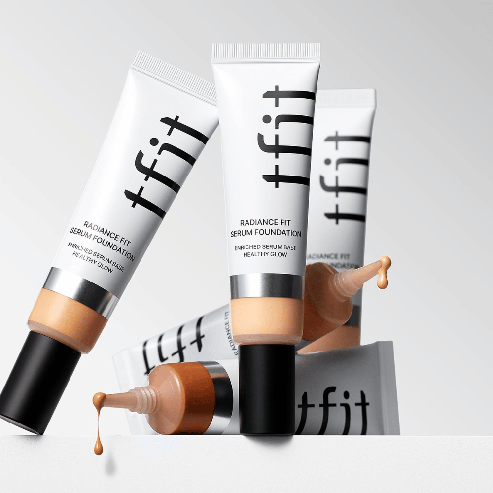 Vários tubos de TFIT Radiance Fit Serum Foundation em diferentes tons, um dos quais com foundation a pingar, contra um fundo cinza claro.
