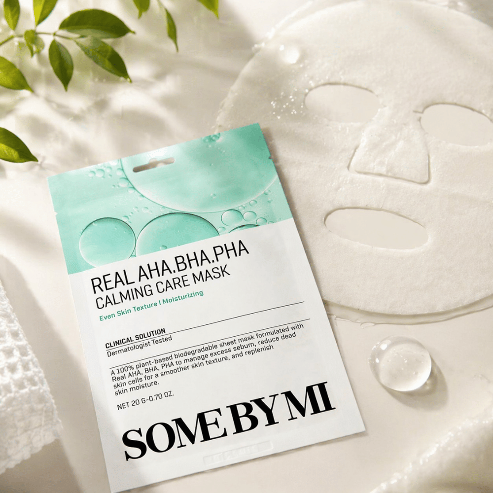 SOME BY MI Real AHA.BHA.PHA Calming Care Mask é apresentada juntamente com a máscara de tecido desdobrada. À volta do produto vêem-se gotas de humidade e folhas verdes que reforçam a sensação de hidratação, cuidado calmante e esfoliação suave. A imagem destaca o foco da máscara numa textura de pele mais uniforme e num melhor equilíbrio de hidratação.