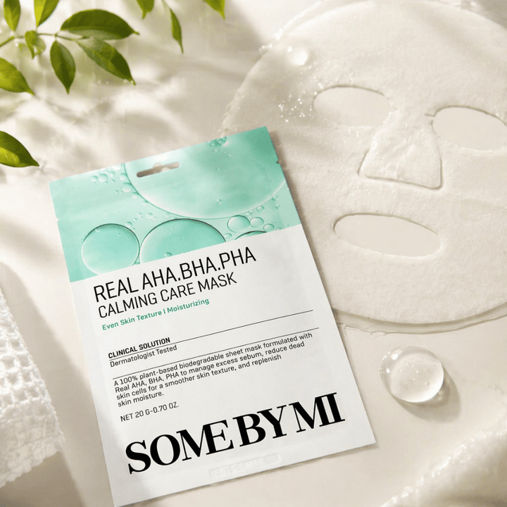 SOME BY MI Real AHA.BHA.PHA Calming Care Mask é apresentada juntamente com a máscara de tecido desdobrada. À volta do produto vêem-se gotas de humidade e folhas verdes que reforçam a sensação de hidratação, cuidado calmante e esfoliação suave. A imagem destaca o foco da máscara numa textura de pele mais uniforme e num melhor equilíbrio de hidratação.