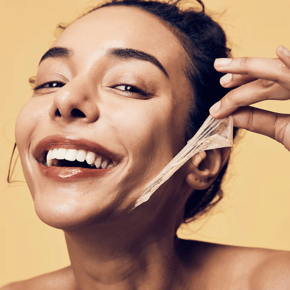 A imagem mostra uma mulher com um grande sorriso removendo uma máscara peel-off do rosto. A mulher parece feliz e satisfeita enquanto usa o produto de beleza, transmitindo uma sensação de bem-estar e cuidado com a pele.