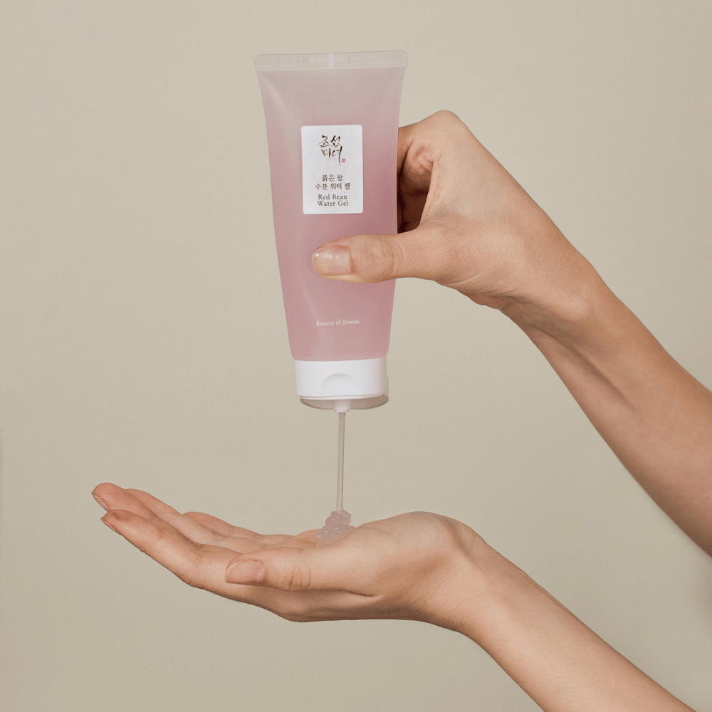 A imagem mostra uma pessoa segurando um tubo de "Red Bean Water Gel" da Beauty of Joseon e espremendo um gel transparente em sua mão. O produto parece ser destinado a cuidados com a pele e tem uma embalagem atraente, de cor rosa suave.