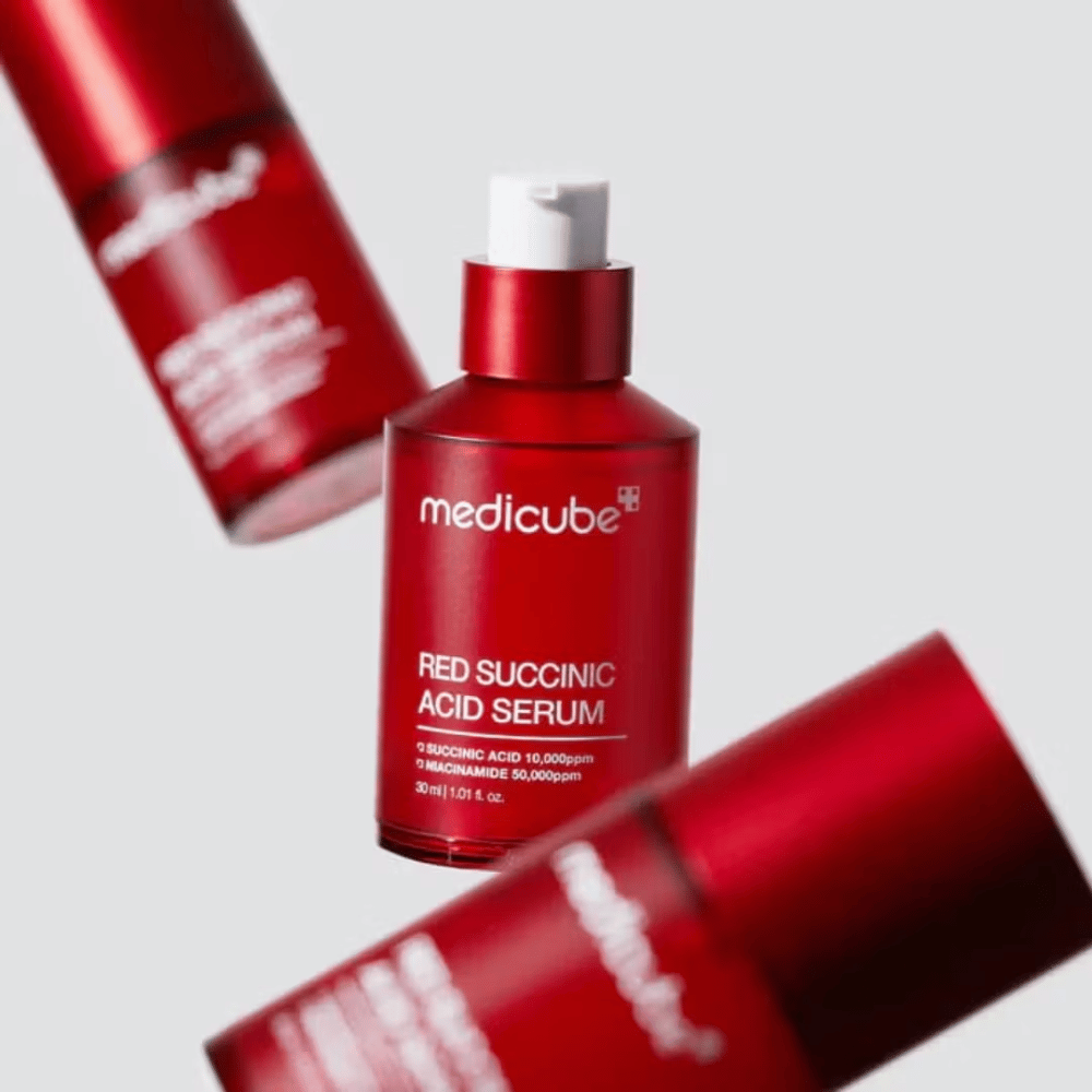 Medicube Red Succinic Acid Serum em frasco de bomba vermelho, cercado por mais frascos idênticos contra fundo claro. O produto contém ácido succínico e niacinamida para tratar acne e manchas de pigmentação.