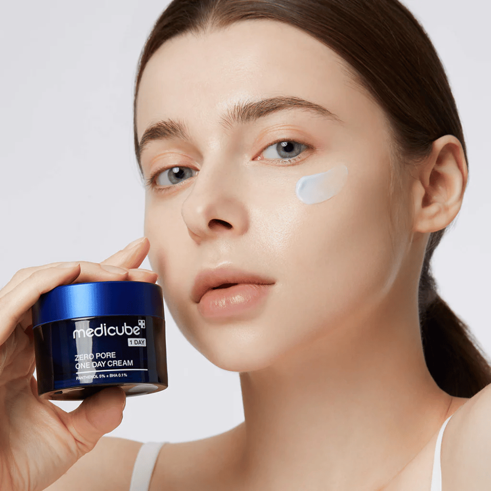 Mulher com um visual natural segura Medicube Zero Pore One Day Cream. Uma pequena quantidade de creme é aplicada em sua bochecha. O produto é projetado para melhorar a textura da pele e minimizar o tamanho dos poros, com uma fórmula leve e hidratante.