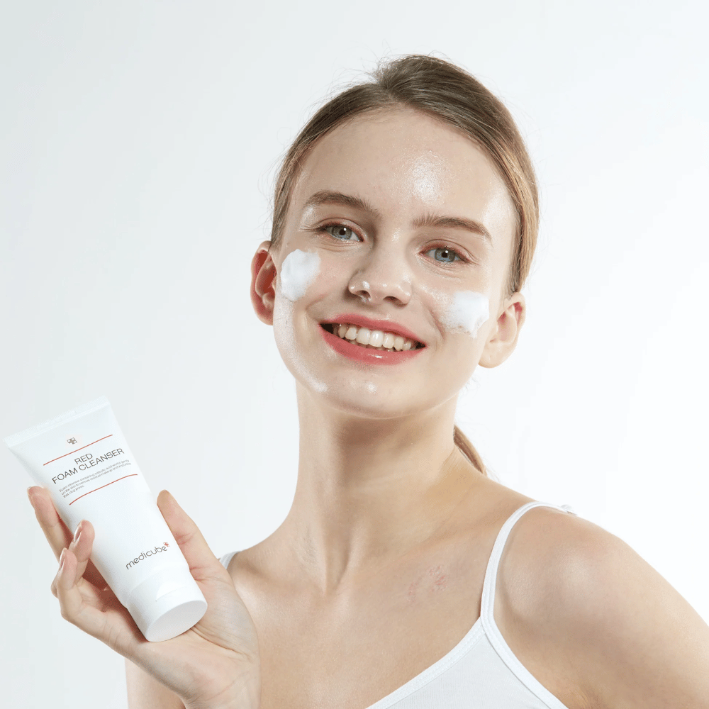 Jovem mulher com espuma de limpeza no rosto segurando um tubo de Medicube Red Foam Cleanser e sorrindo para a câmera. Ela está usando uma camisola branca e o fundo é claro e simples.