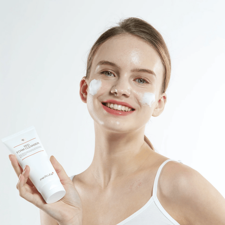 Ung kvinna med rengöringsskum i ansiktet håller upp en tub av Medicube Red Foam Cleanser och ler mot kameran. Hon bär ett vitt linne och bakgrunden är ljus och enkel.