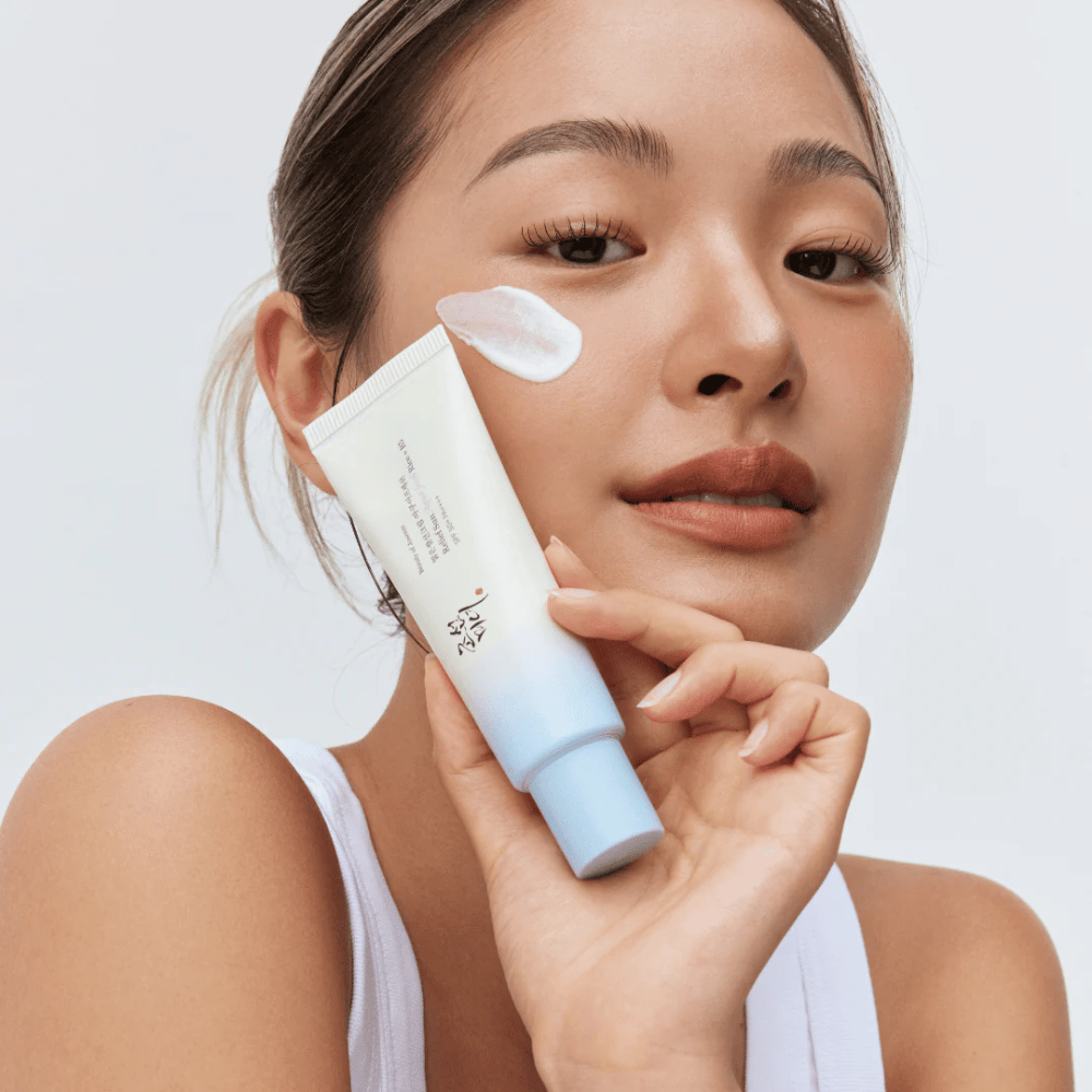 Mulher segura Beauty of Joseon Relief Sun: protetor solar Aqua-Fresh Rice + B5 e mostra sua consistência cremosa na bochecha. Um produto leve de proteção solar que hidrata e protege a pele com SPF 50+ PA++++.