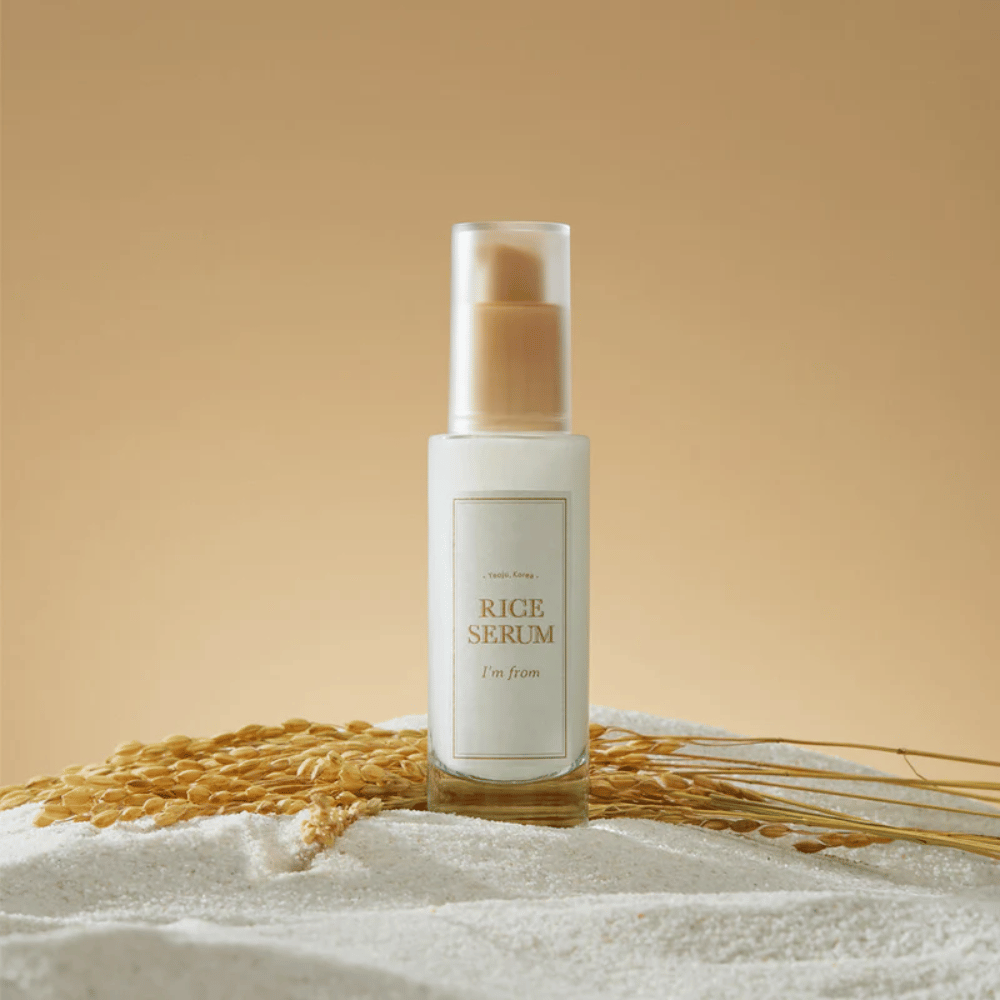 Garrafa com "I'm from" Rice Serum, cercada por grãos de arroz e espigas de arroz contra um fundo quente.