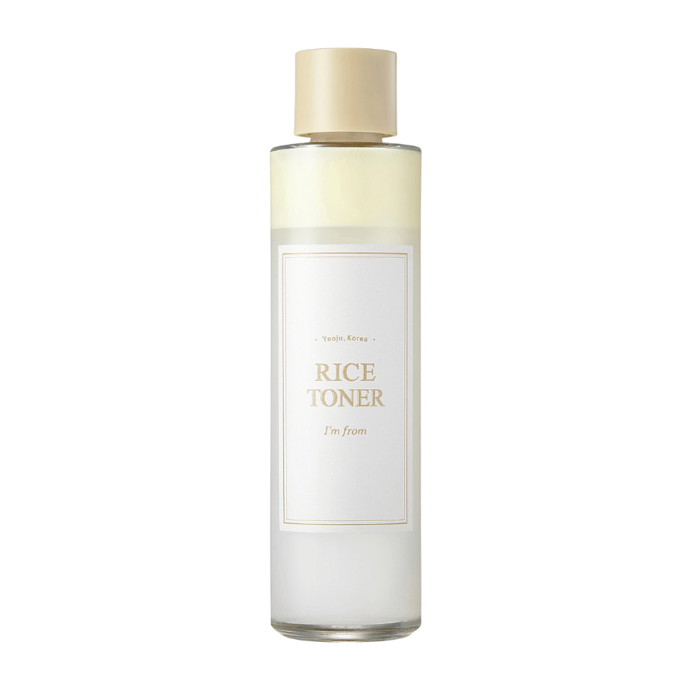 Uma garrafa transparente com tampa dourada e rótulo que indica "Rice Toner I'm from Yeojü, Coreia". A garrafa contém um líquido claro, provavelmente um toner à base de extrato de arroz. O design é limpo e minimalista. O fundo é branco.