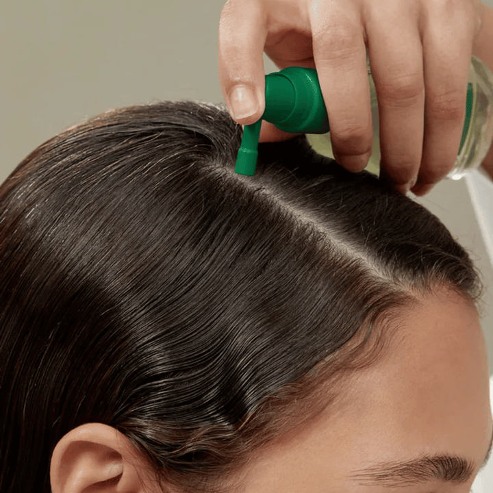 Pessoa aplica AROMATICA Rosemary Root Enhancer diretamente no couro cabeludo com a ajuda do bico direcionado. A imagem mostra como o produto é utilizado para o cuidado do couro cabeludo e o fortalecimento da raiz do cabelo.