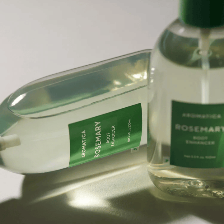 Dois frascos de AROMATICA Rosemary Root Enhancer com tampa protetora transparente e rótulo verde. Grande plano que destaca o design premium do produto e o conceito de cuidados capilares naturais.