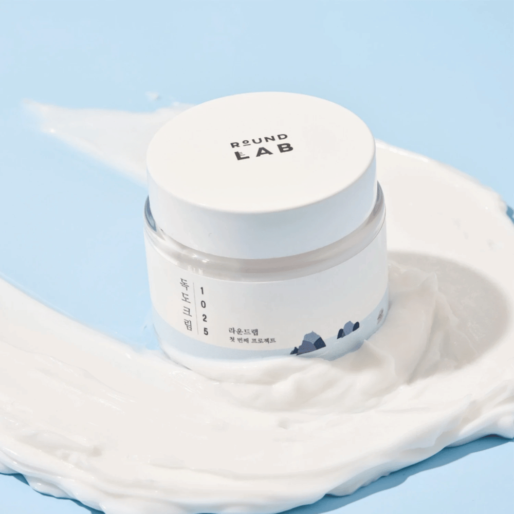 Um frasco de Round Labs 1025 Dokdo Cream 80 ml que proporciona hidratação, colocado sobre um fundo azul vibrante.