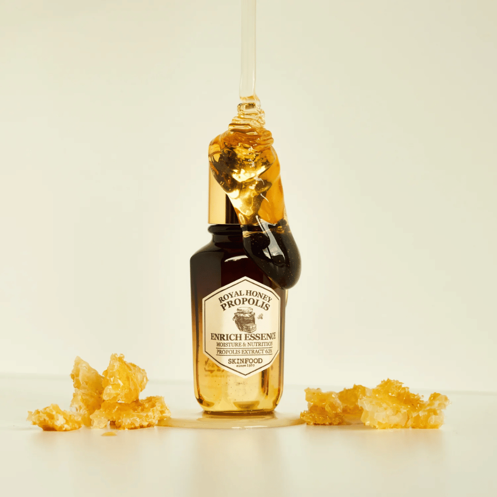 Imagem de uma garrafa de vidro com Royal Honey Propolis Enrich Essence de Skinfood. Uma grande gota de mel dourado escorre da tampa dourada da garrafa. Ao redor da garrafa estão espalhados pequenos pedaços de favo de mel sobre uma superfície clara. A essência é comercializada com foco em umidade e nutrição, com 63% de extrato de própolis.
