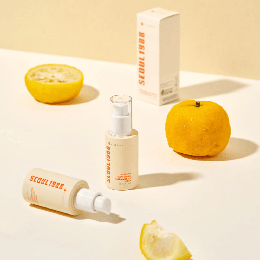 Vários frascos e embalagens de Seoul 1988 Glow Serum arranjados com frutas de yuja inteiras e cortadas ao fundo.