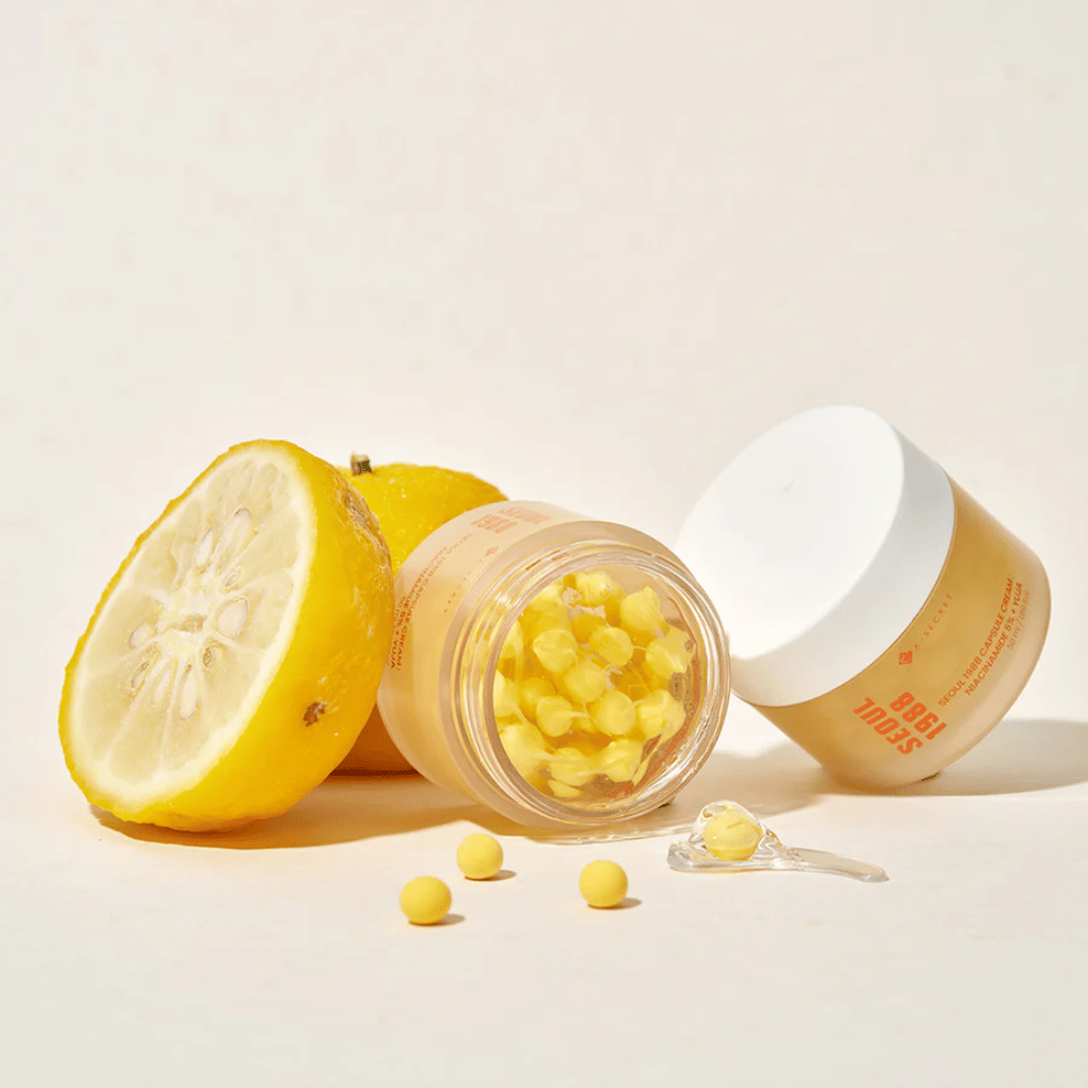 Frasco aberto com cápsulas amarelas ao lado de uma fruta yuja cortada ao meio que destaca a fórmula rica em vitamina C.