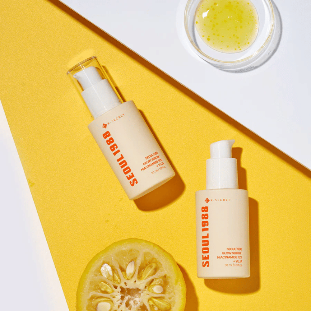 Dois frascos de Seoul 1988 Glow Serum colocados sobre um fundo amarelo junto com uma tigela de soro amarelo e uma fruta cítrica cortada ao meio.