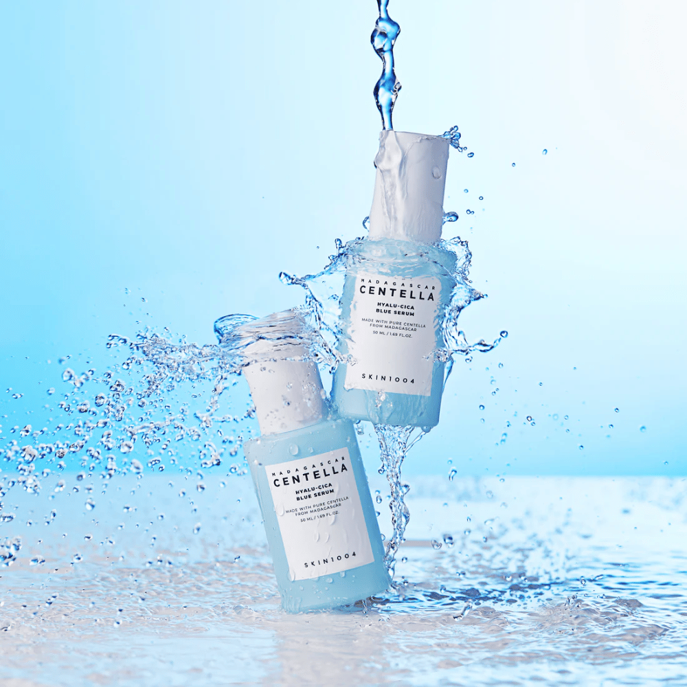 Duas garrafas de SKIN1004 Madagascar Centella Hyalu-Cica Blue Serum são atingidas por uma cascata de água contra um fundo azul claro.