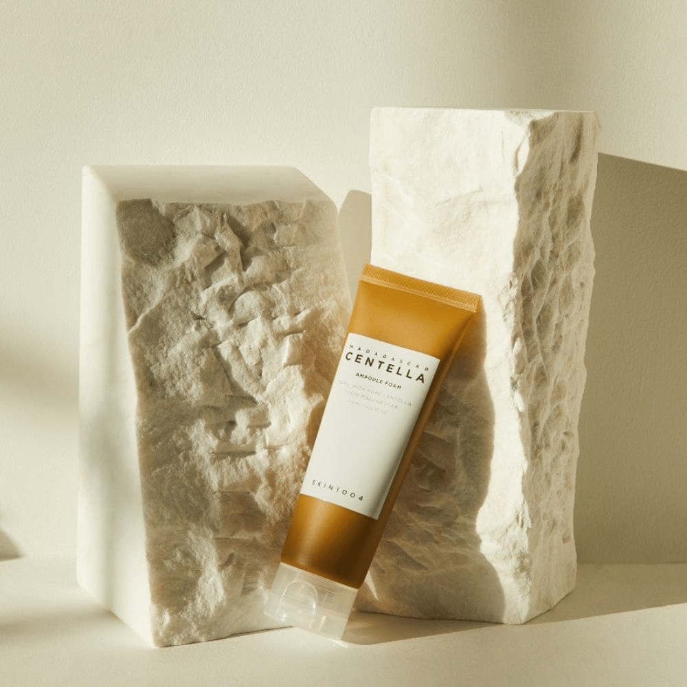 SKIN1004 Madagascar Centella Ampoule Foam de pé contra blocos de pedra clara em luz solar natural, com seu design de tubo marrom e branco e minimalismo elegante.