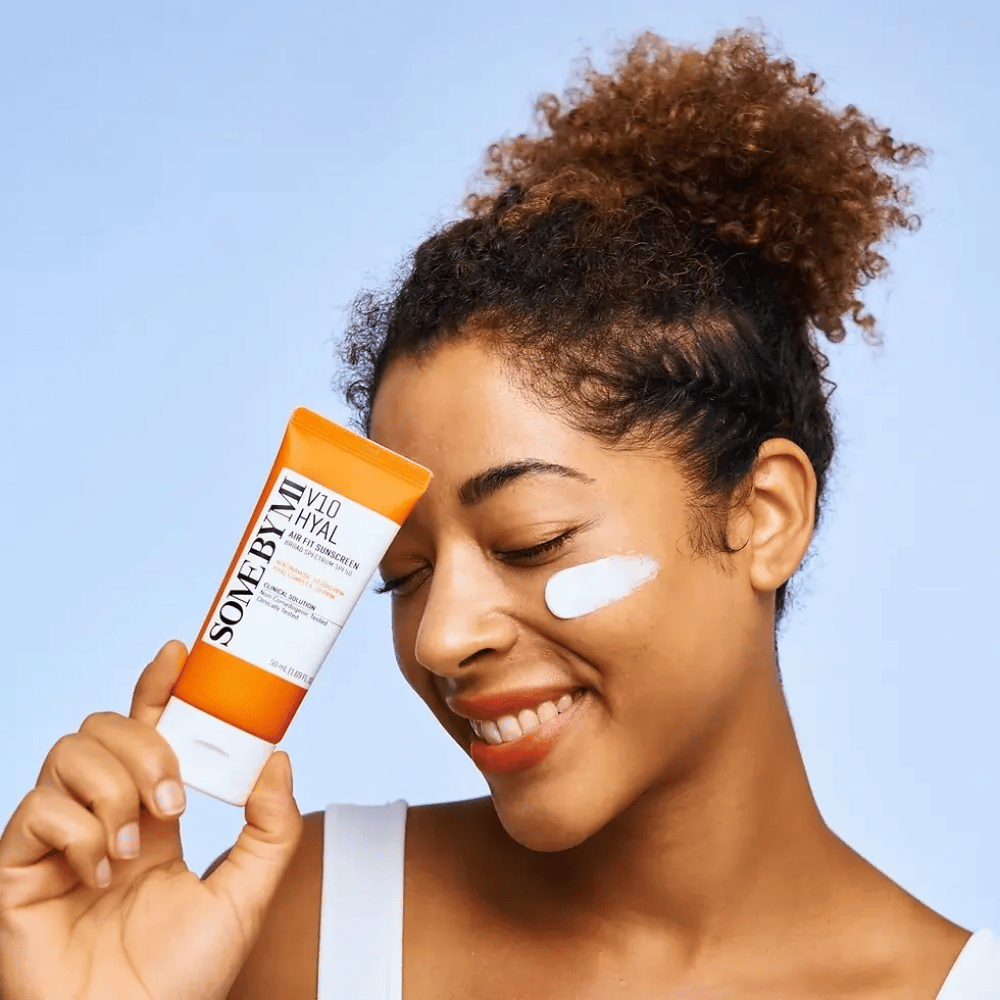 Uma mulher segura um tubo de SOME BY MI V10 Hyal Air Fit Sunscreen e tem uma mancha branca de protetor solar na bochecha. Ela sorri alegremente e destaca claramente o produto, enfatizando seu uso e benefícios para os cuidados com a pele e proteção solar.
