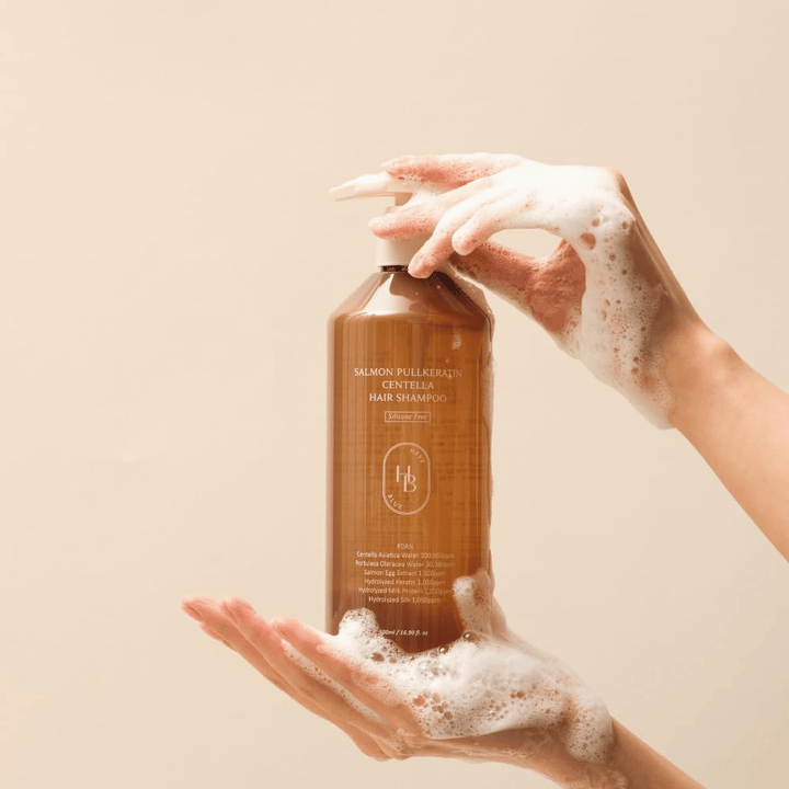 Heveblue Salmon Pullkeratin Centella Hair Shampoo em frasco castanho rodeado de espuma. O produto é segurado entre duas mãos e destaca uma experiência de cuidado capilar de limpeza e nutrição com espuma abundante.