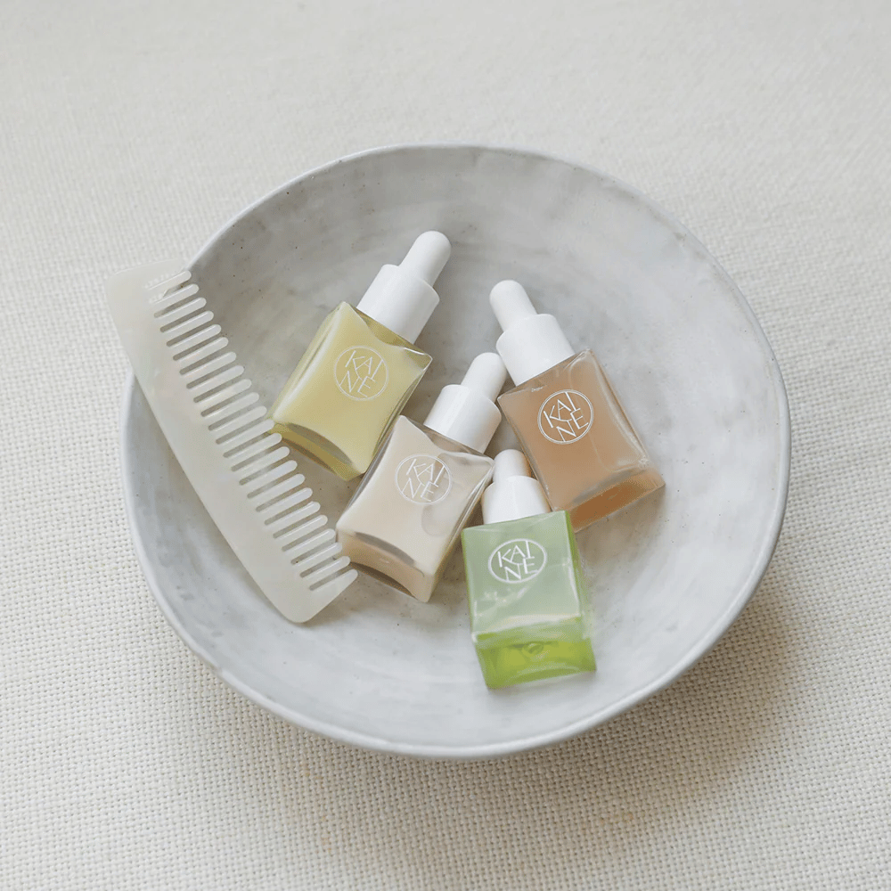 Quatro pequenos frascos de soro da KAINÉ Serum Discovery Kit estão em uma tigela branca ao lado de um pente, com um fundo natural e calmo em tom bege.