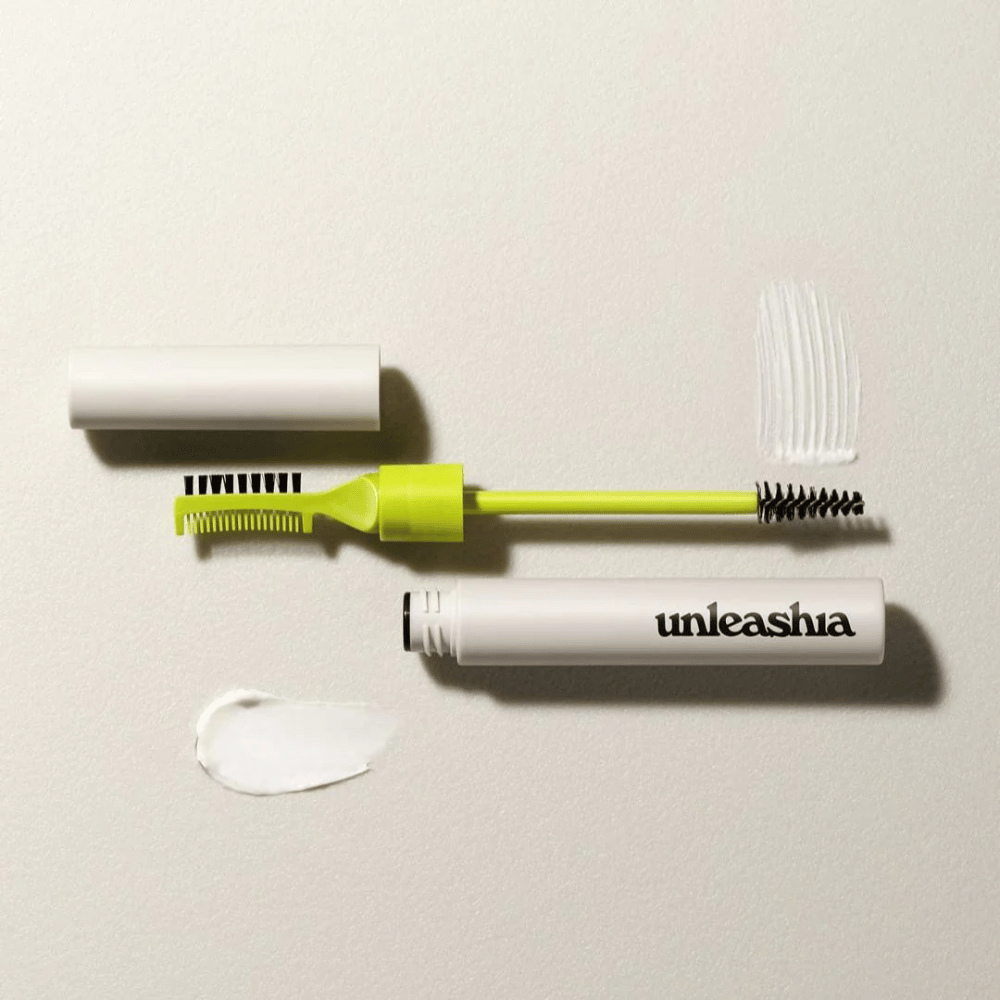 Um conjunto de escultura de sobrancelhas com um pente verde, escova preta e um tubo branco marcado "unleashia" sobre um fundo claro com um montinho de produto transparente.