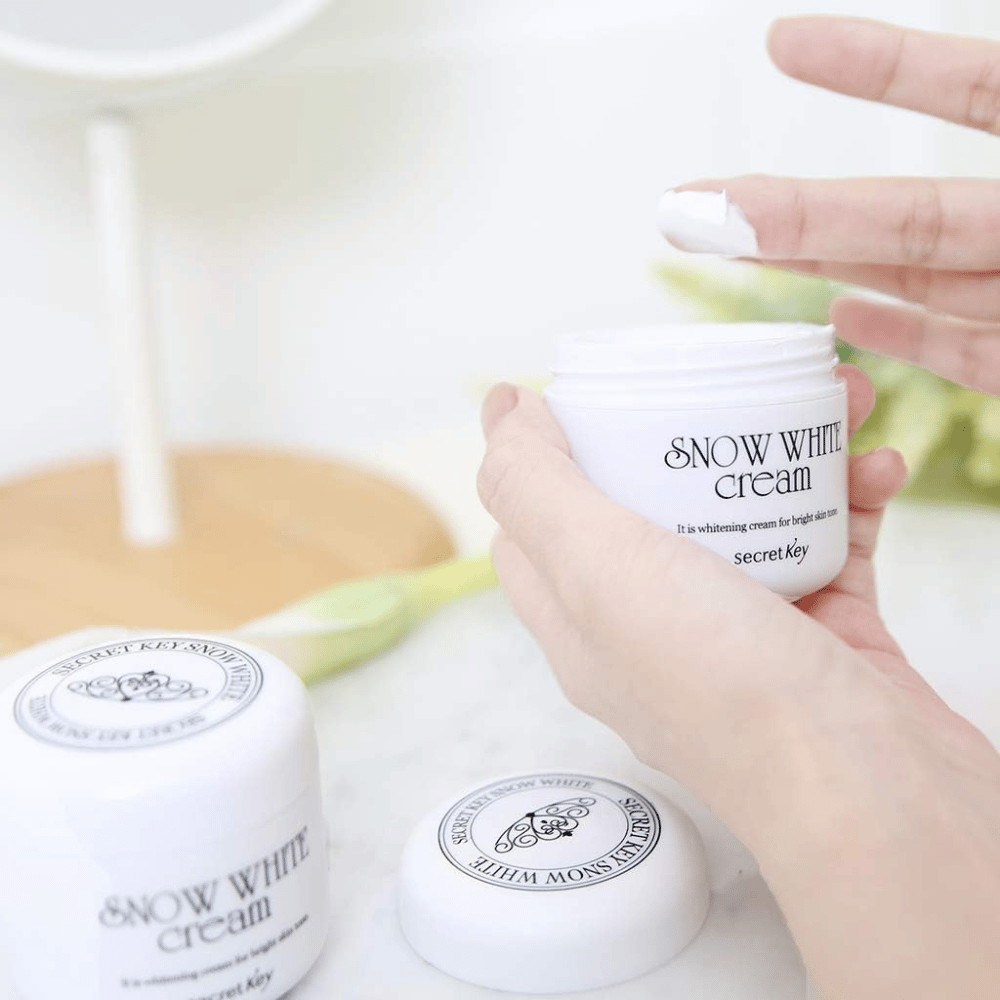 Mão pegando creme branco de um pote Secret Key Snow White Cream em uma penteadeira.