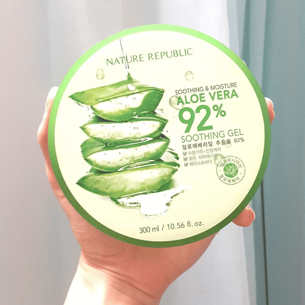 Uma mão segura uma lata redonda com Nature Republic Aloe Vera 92% Gel Suavizante. A lata é verde com uma imagem de aloe vera e texto que descreve suas propriedades calmantes e hidratantes. O conteúdo é de 300 ml.