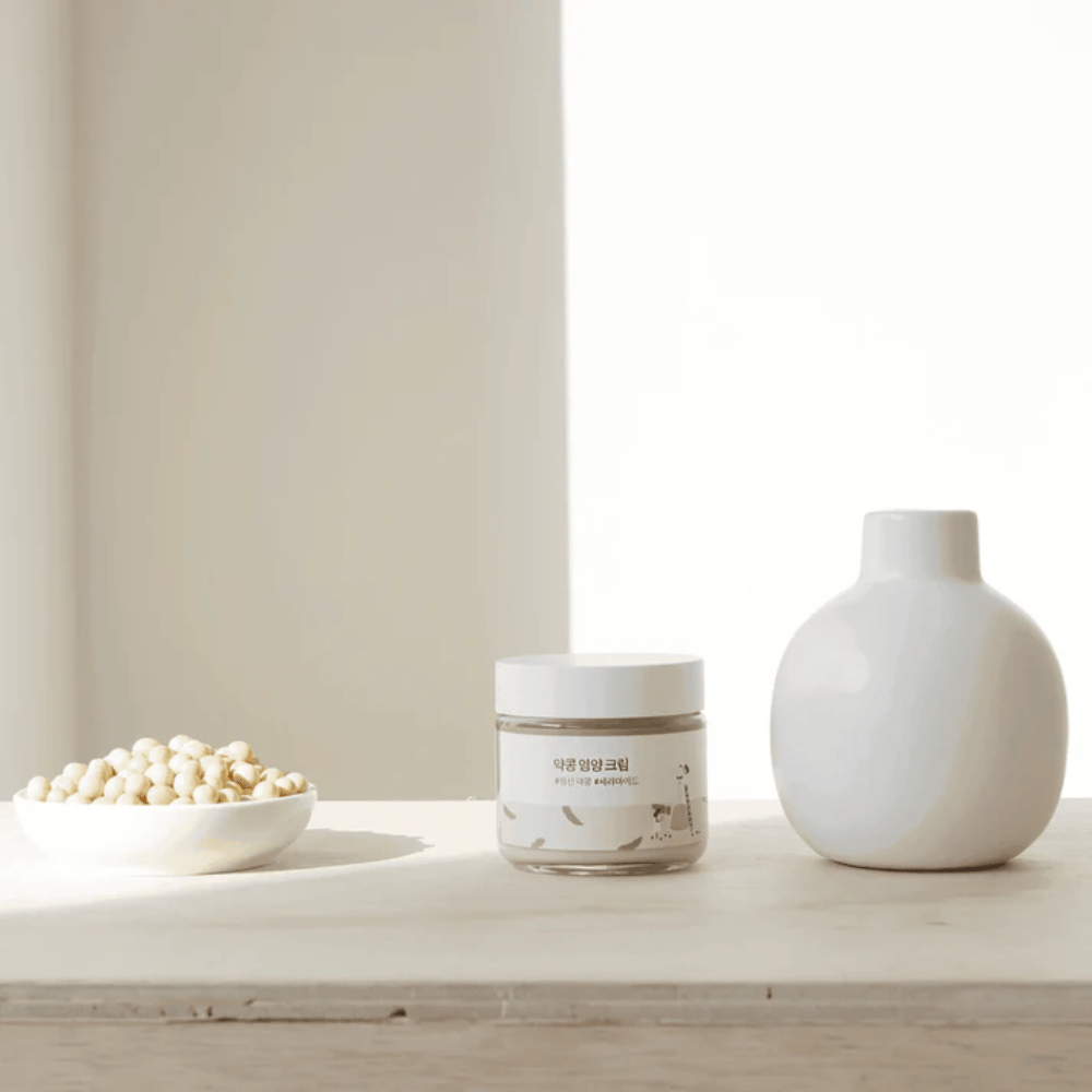 Mixsoon Soybean Nourishing Cream está sobre uma mesa de madeira ao lado de um vaso e de grãos de soja. Um creme minimalista e hidratante com ingredientes naturais.
