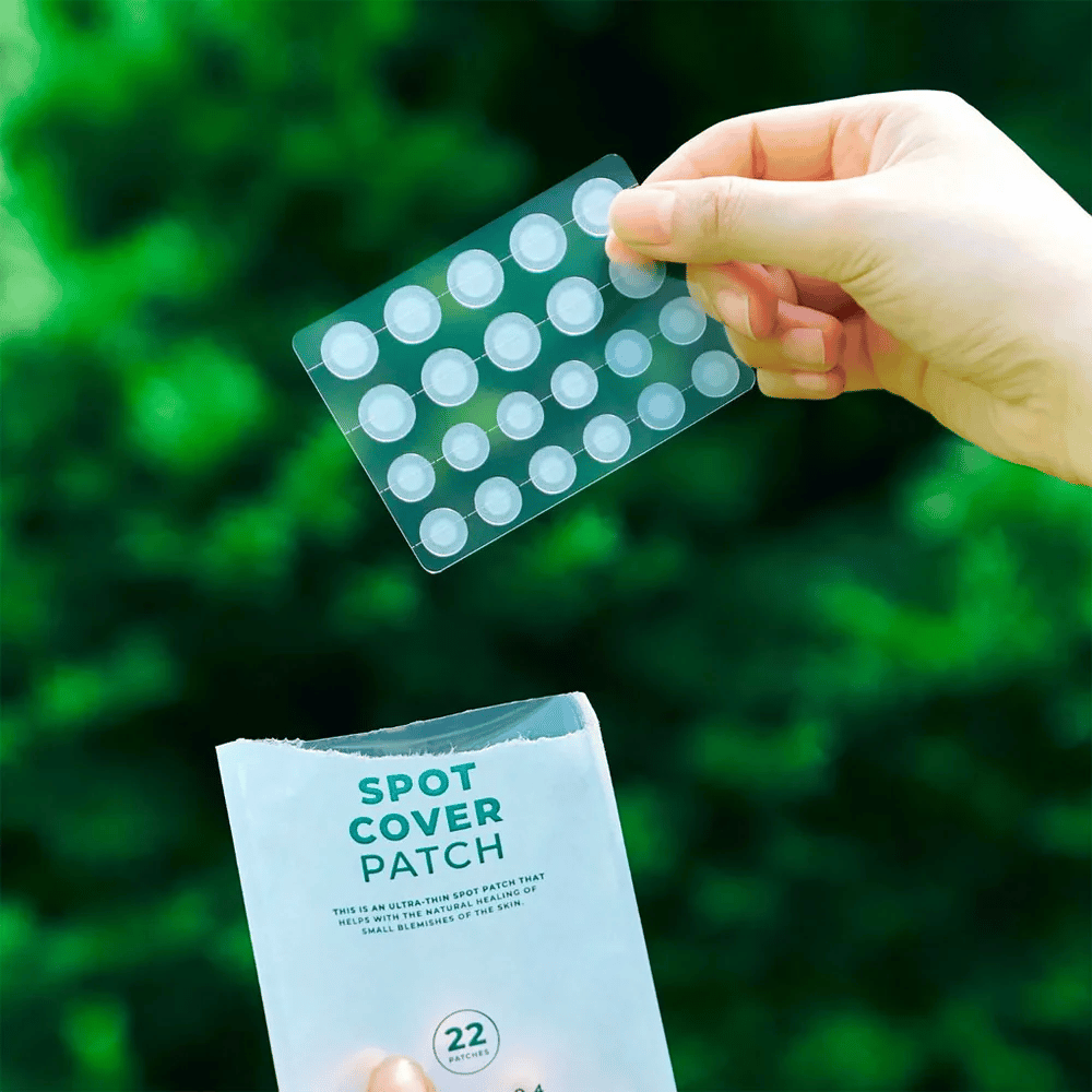 Uma mão segura uma folha de Spot Cover Patches, que são transparentes e arredondados. Ao fundo, vê-se um ambiente verde desfocado. A embalagem mostra uma parte da embalagem com o texto "SPOT COVER PATCH" e informações de que o produto contém 22 adesivos que ajudam a curar pequenas áreas da pele.