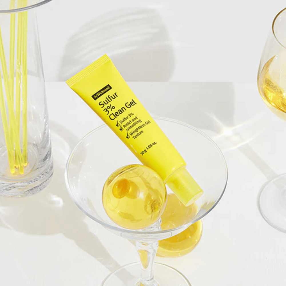 By Wishtrend Sulfur 3% Clean Gel em um tubo amarelo colocado em um copo de cocktail com uma esfera amarela, cercado por elegantes copos em um fundo branco.