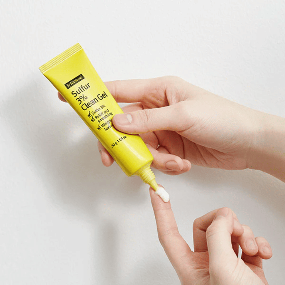 Close-up de mãos aplicando By Wishtrend Sulfur 3% Clean Gel, um tubo amarelo com gel branco, contra um fundo branco.